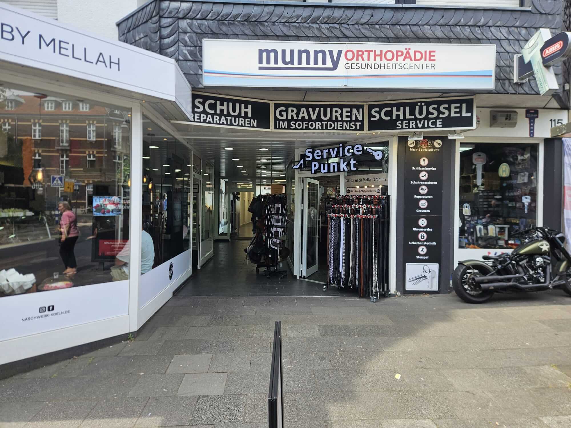 Storefront with signage reading 'munny Orthopädie Gesundheitscenter' and adjacent black panels advertising shoe repair, engraving and key service.